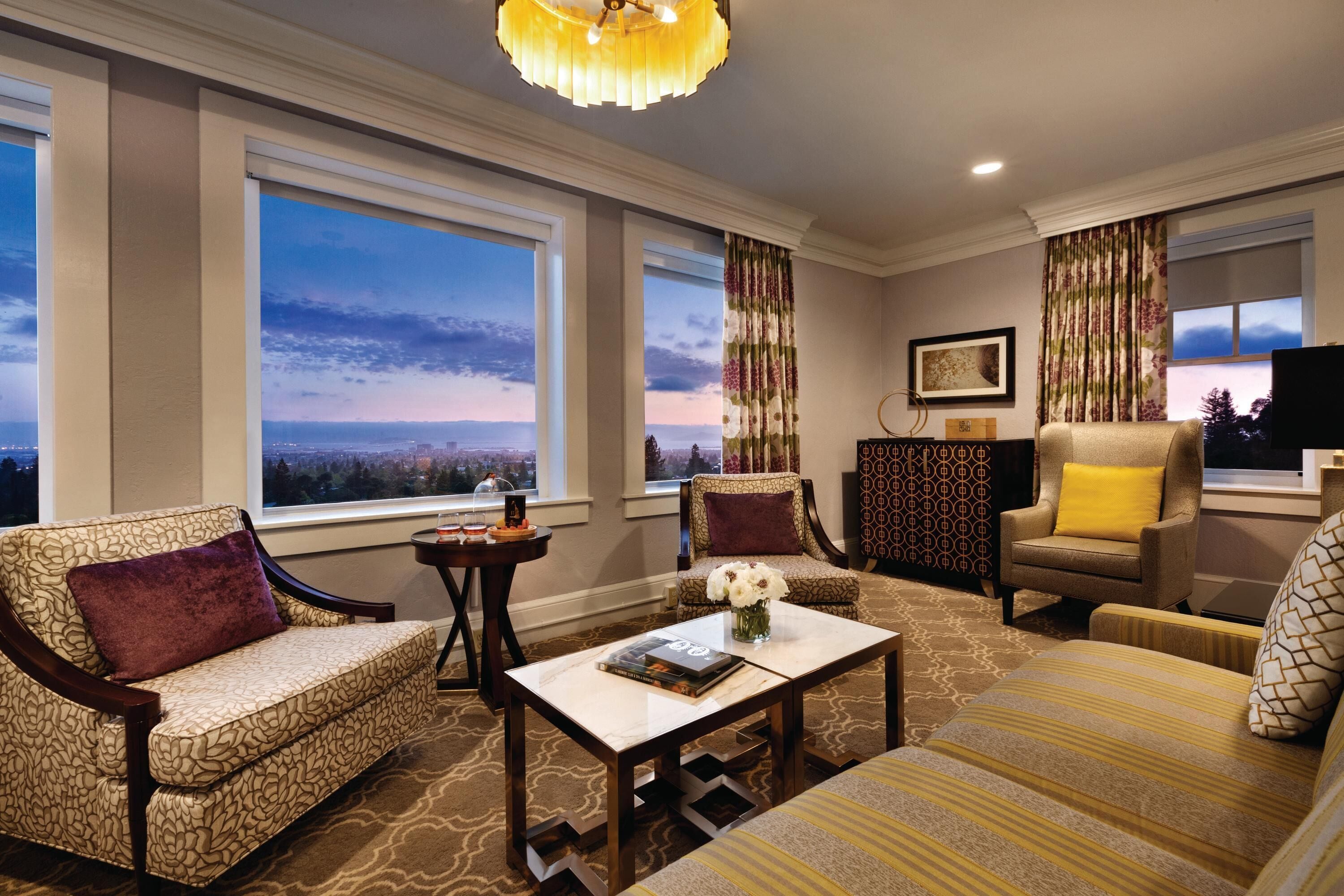 A living room with a sofa, armchairs, a coffee table, and a TV. Large windows offer a view of the cityscape.
