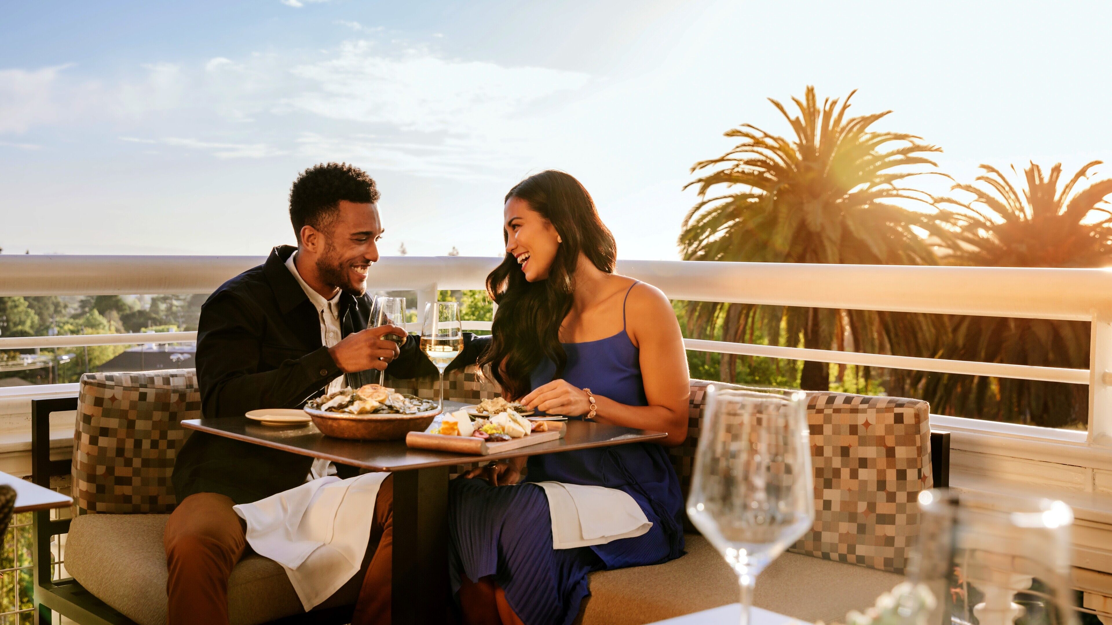 A couple dining at an outdoor table with a view of palm trees and a sunset.