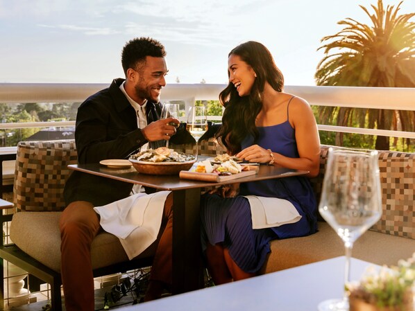 Una pareja cenando al aire libre en un restaurante con vistas a palmeras y al atardecer.