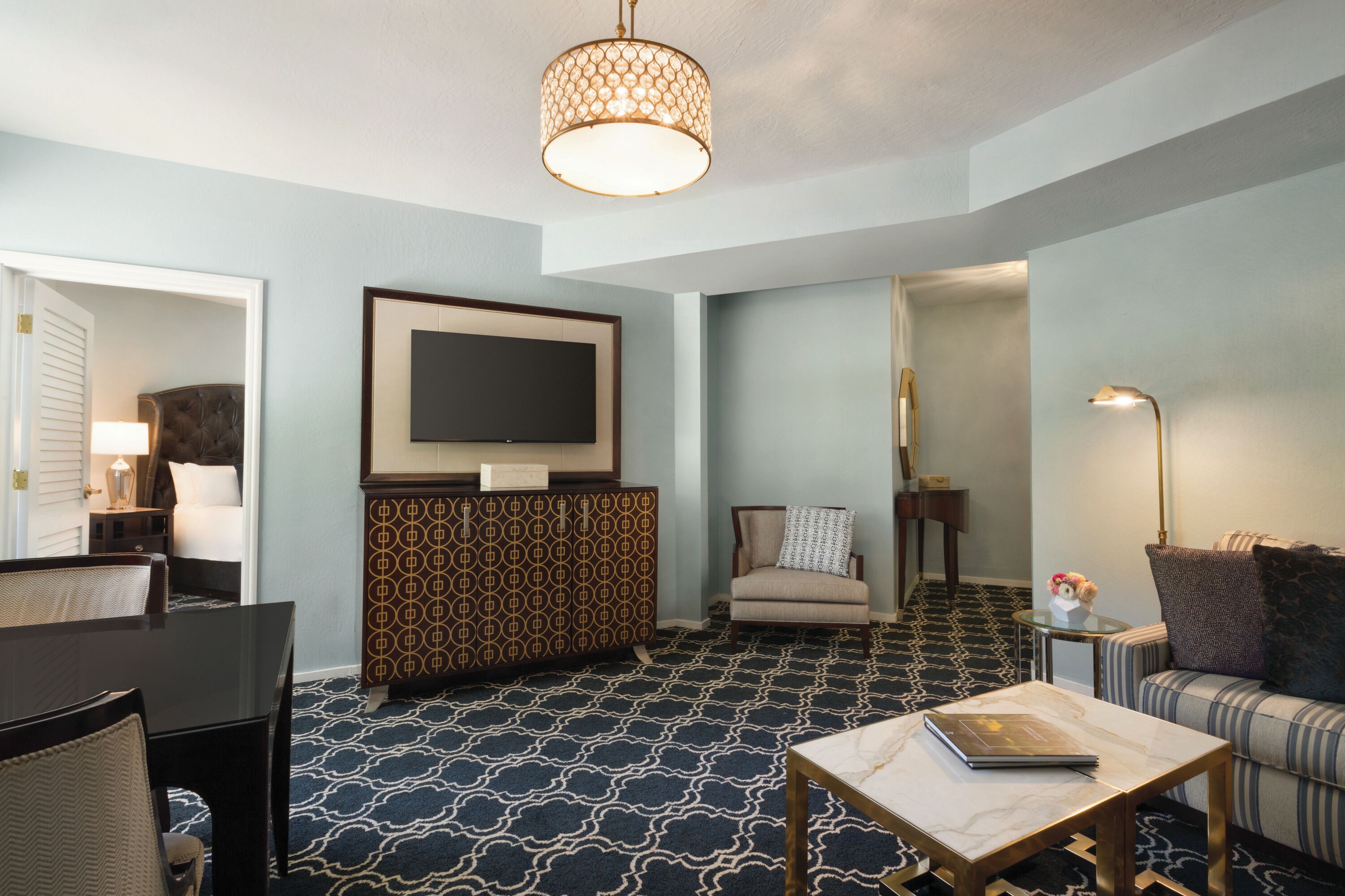 A hotel room with a patterned carpet, a sofa, a coffee table, a television mounted on a cabinet, and a chandelier.