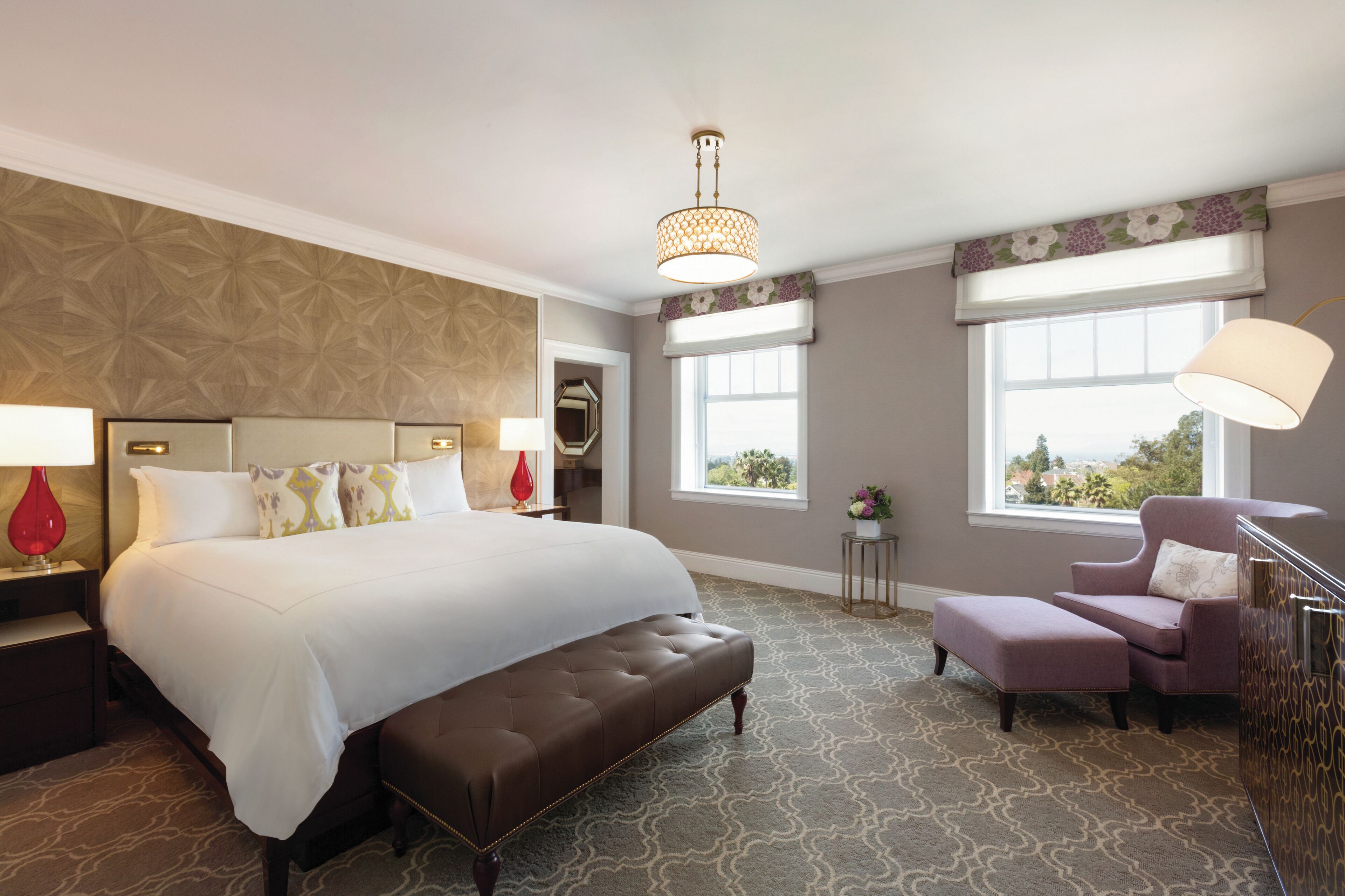 A hotel room with a large bed, a sitting area, a window with a view, and a patterned carpet.