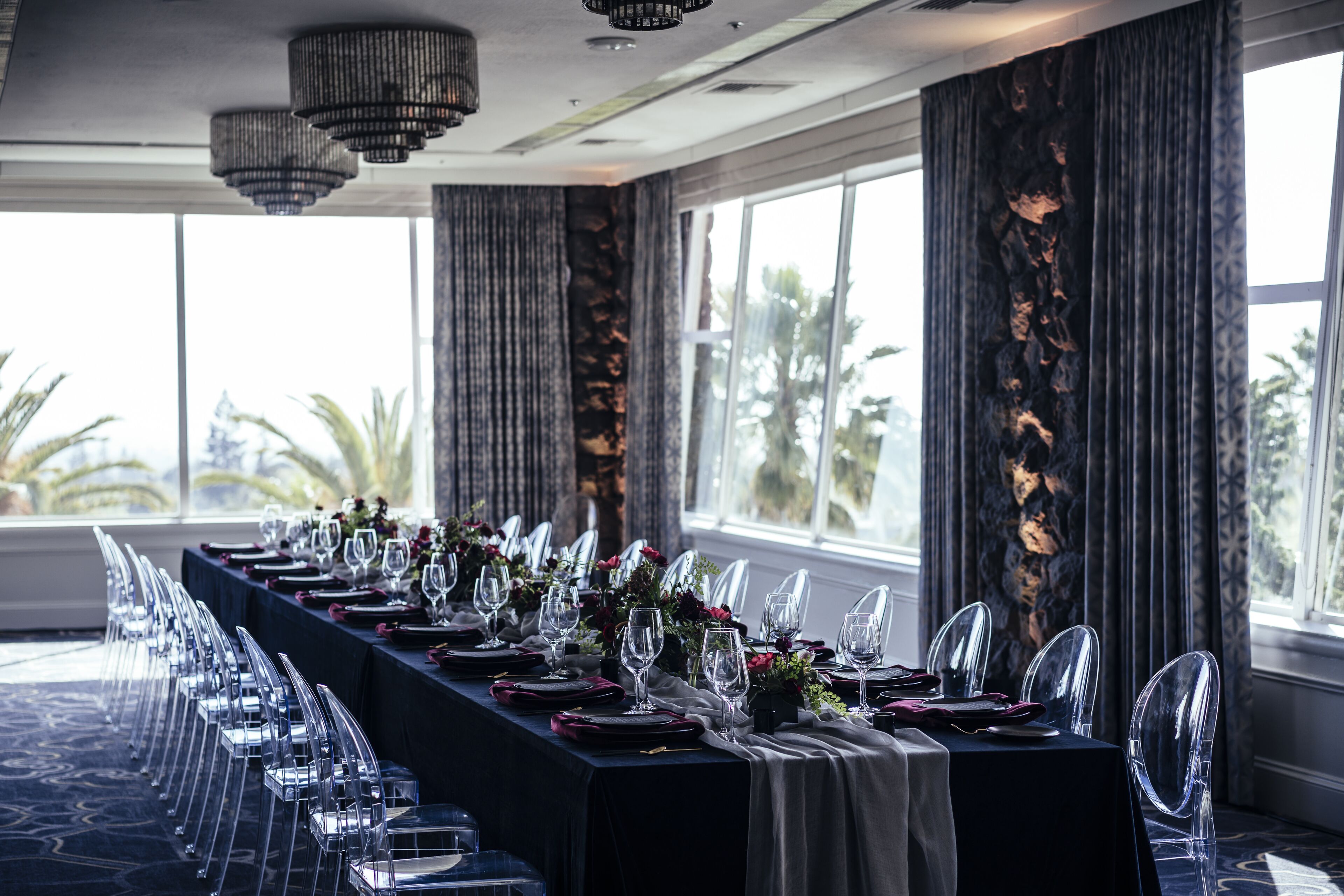 A long dining table set with clear chairs, wine glasses, and plates, with a view of palm trees through large windows.