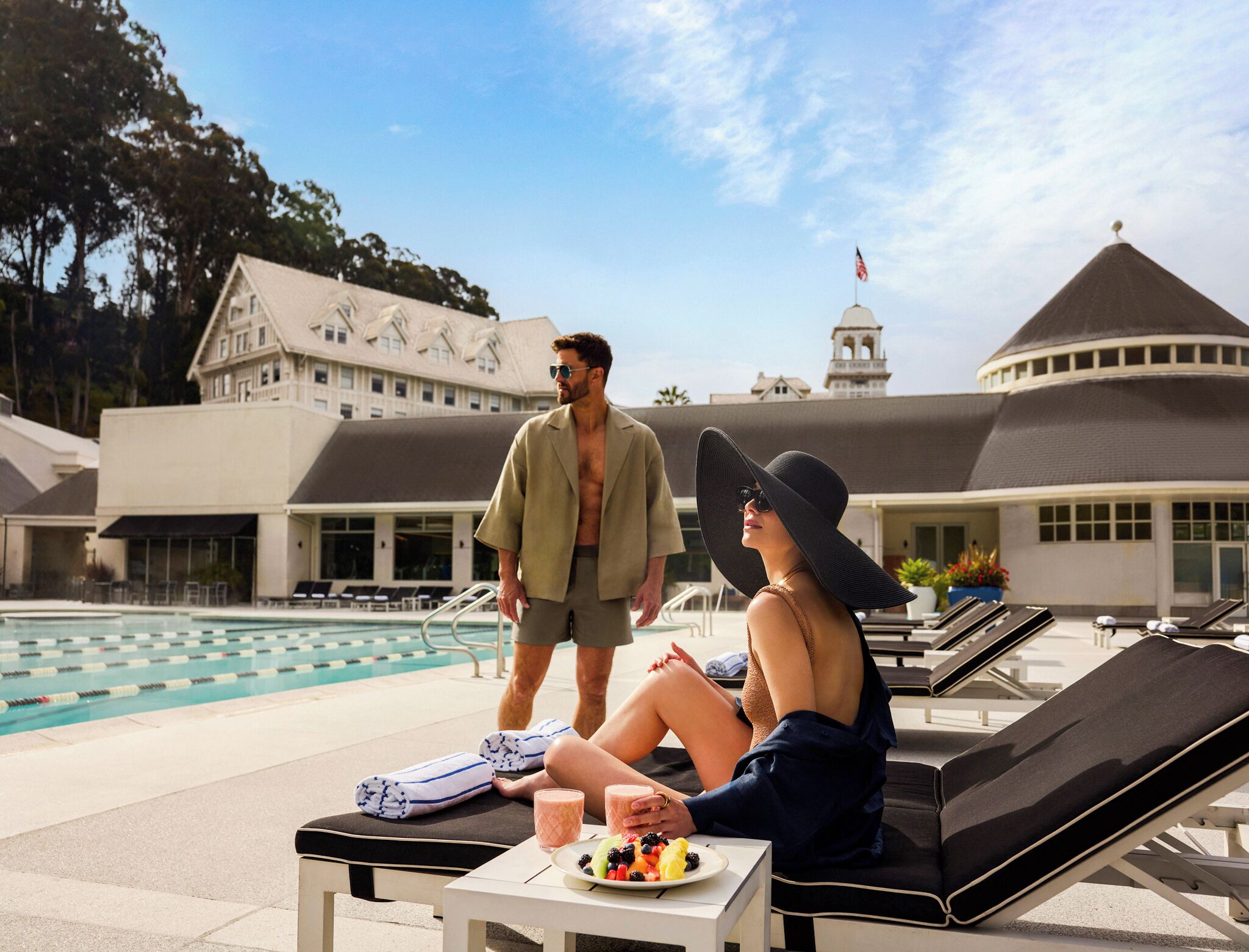 Zona junto a la piscina con sillones, una mesa con una fuente de fruta y un edificio con una torre de reloj al fondo.