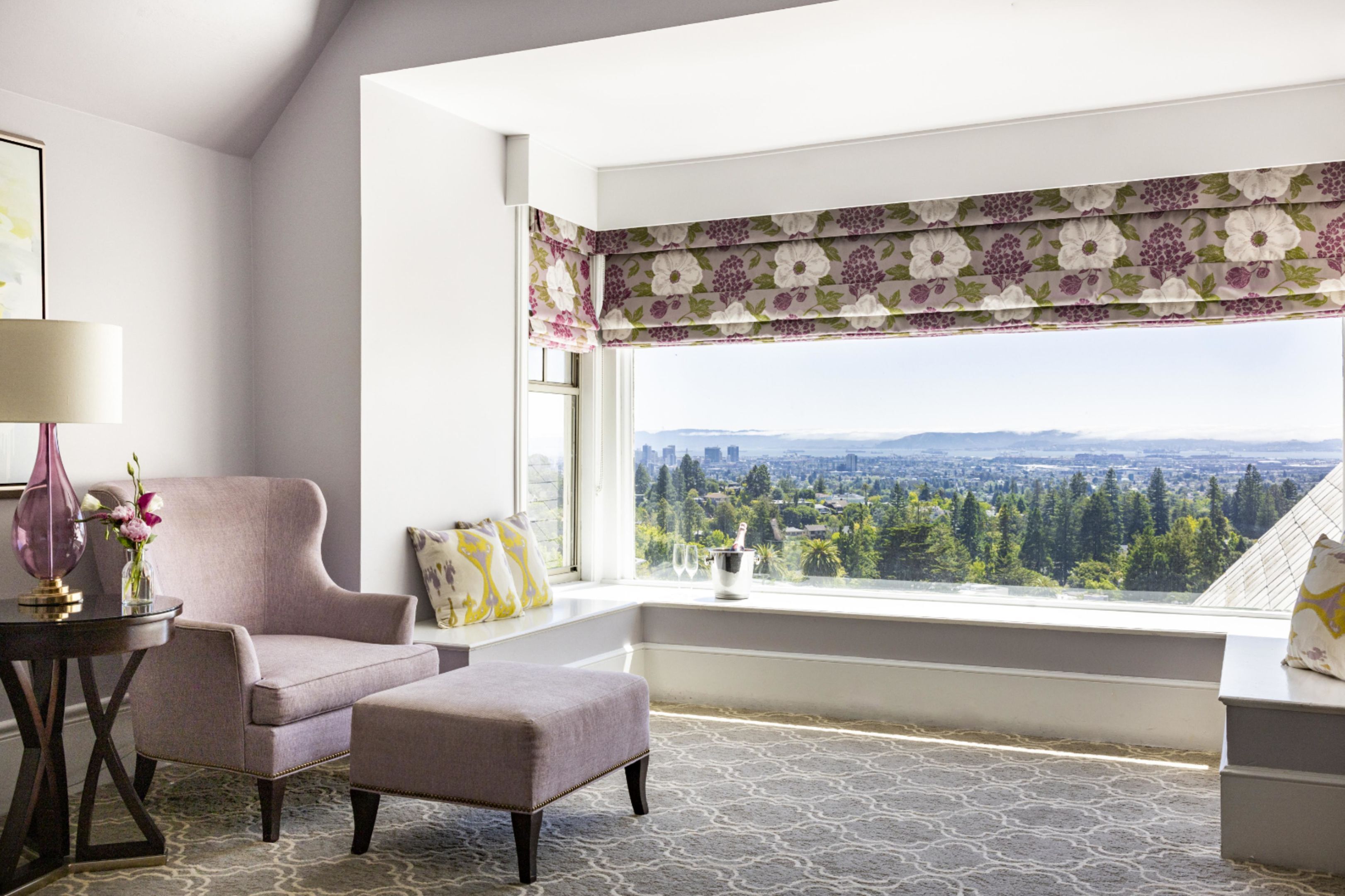 A room with a view of a cityscape, a window with floral curtains, a cushioned chair, and a small table with a vase of flowers.