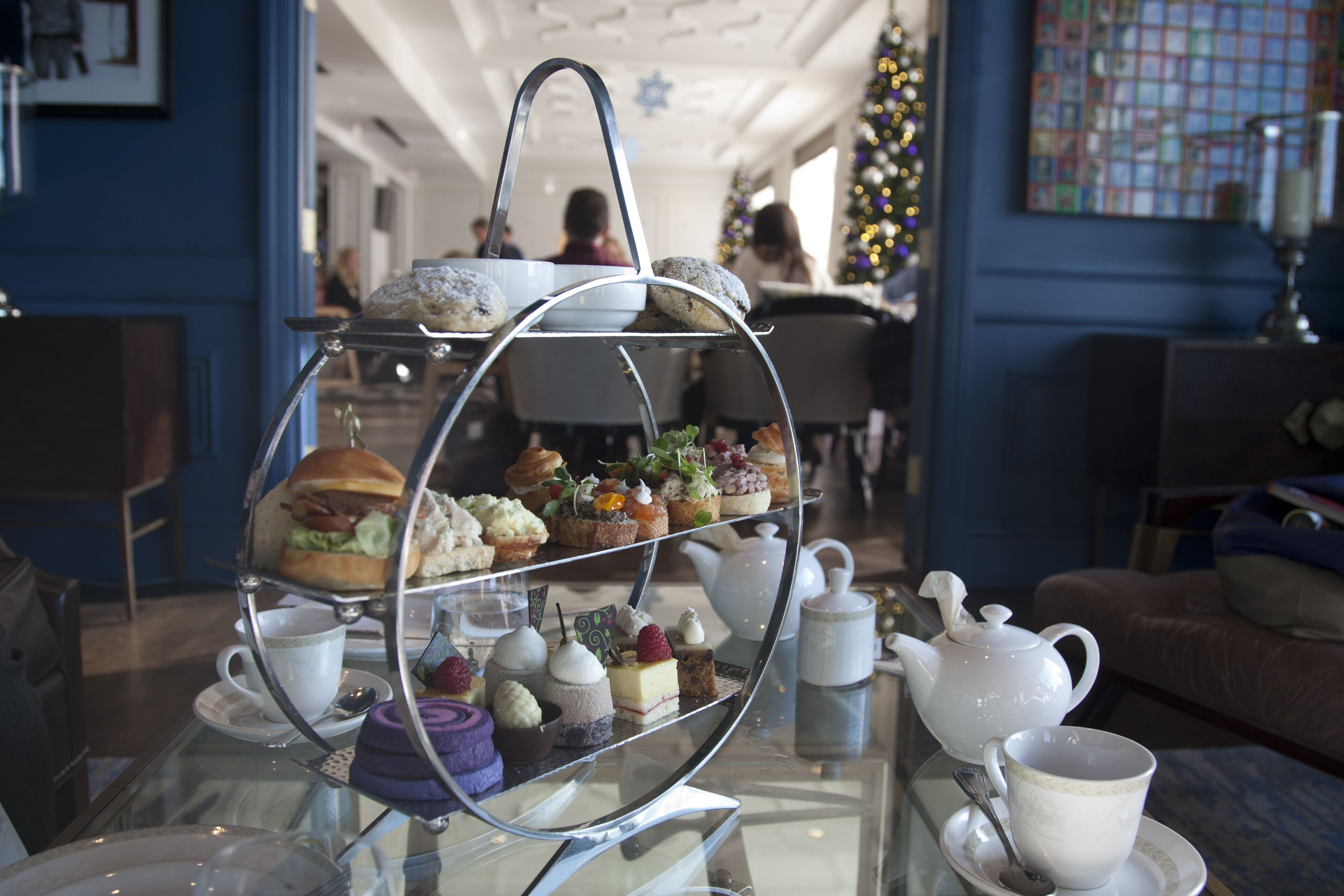 A three-tiered dessert stand with various pastries and a teapot on a glass table.