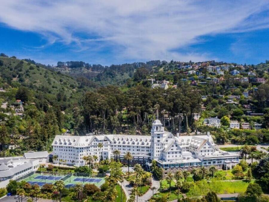 A large, white hotel with multiple wings and a central tower, surrounded by lush greenery and hills.