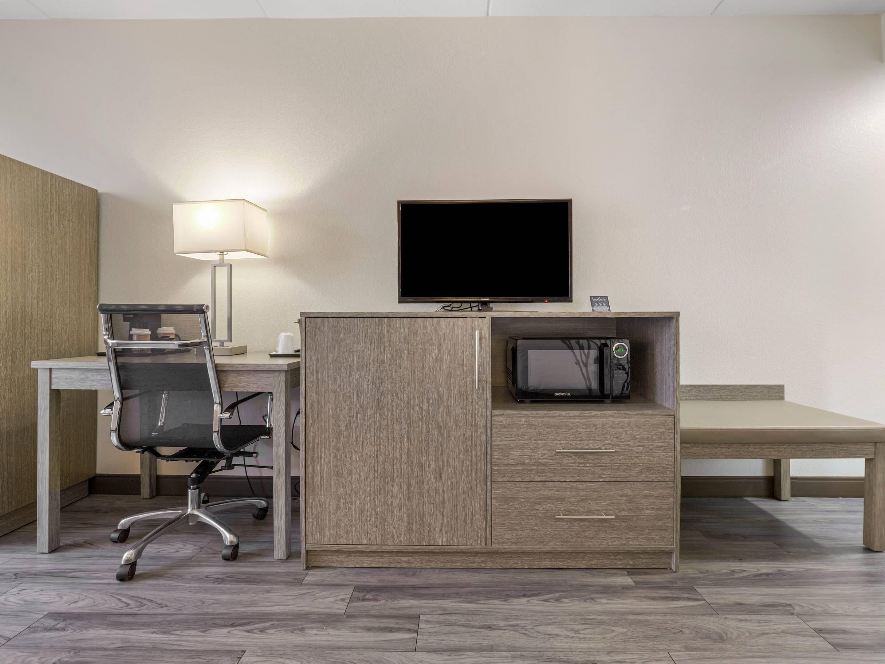 A hotel room with a desk, chair, television, microwave, and a bedside table.