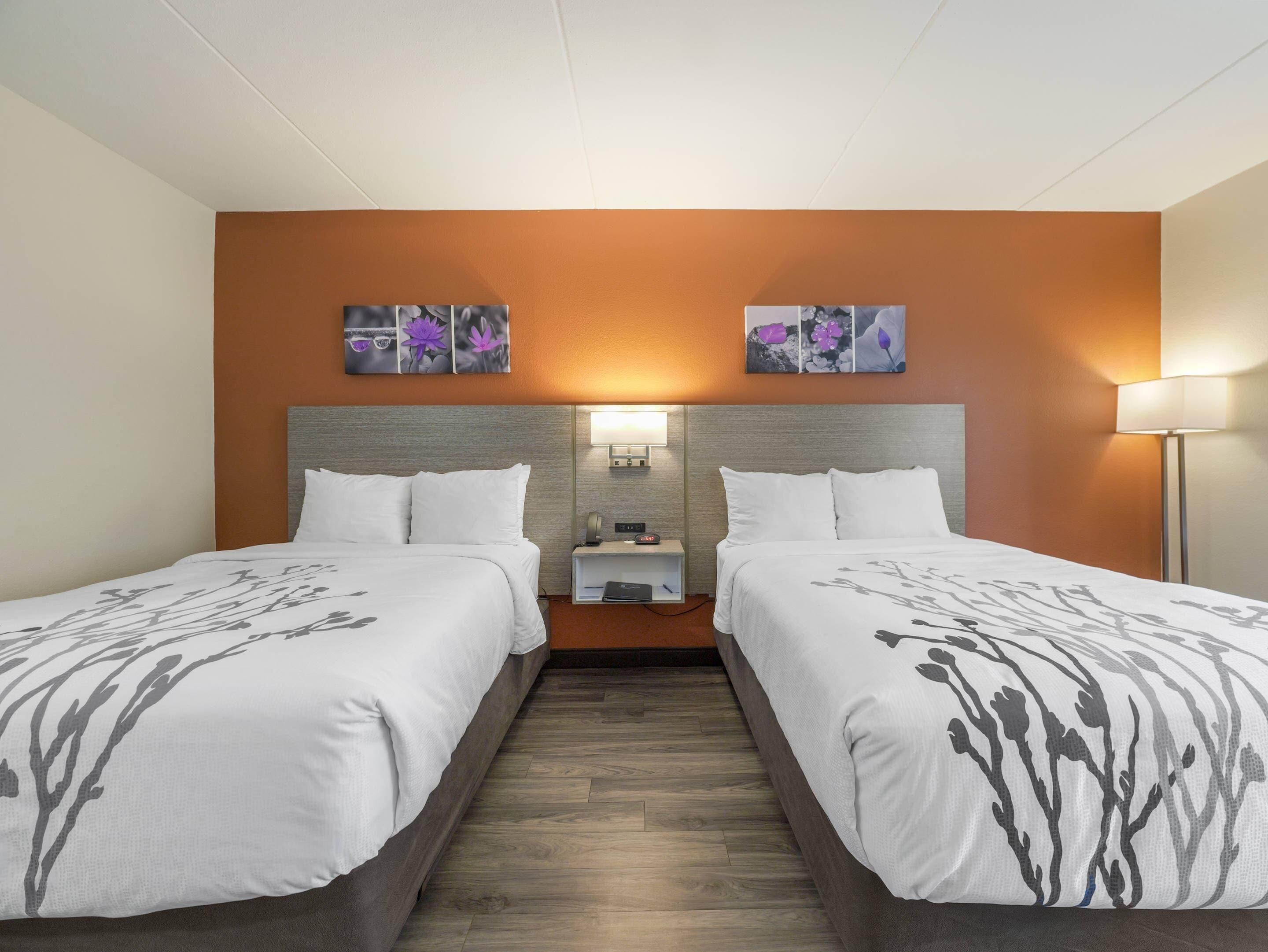A hotel room with two beds, a nightstand, a lamp, and artwork on the wall.