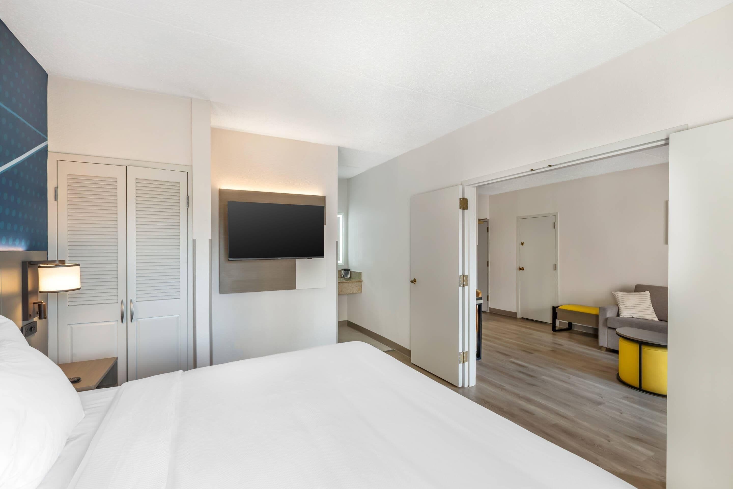 Ein Hotelzimmer mit einem Bett, einem an der Wand montierten Fernseher, einem Nachttisch mit Lampe, einem Kleiderschrank und einer Tür zu einem Wohnbereich mit Sofa und Couchtisch.