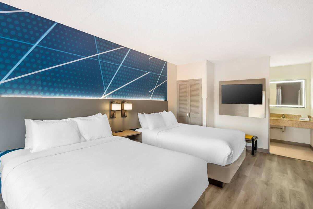 Habitación de hotel con dos camas, un televisor, un baño con espejo y una pared con diseño geométrico.