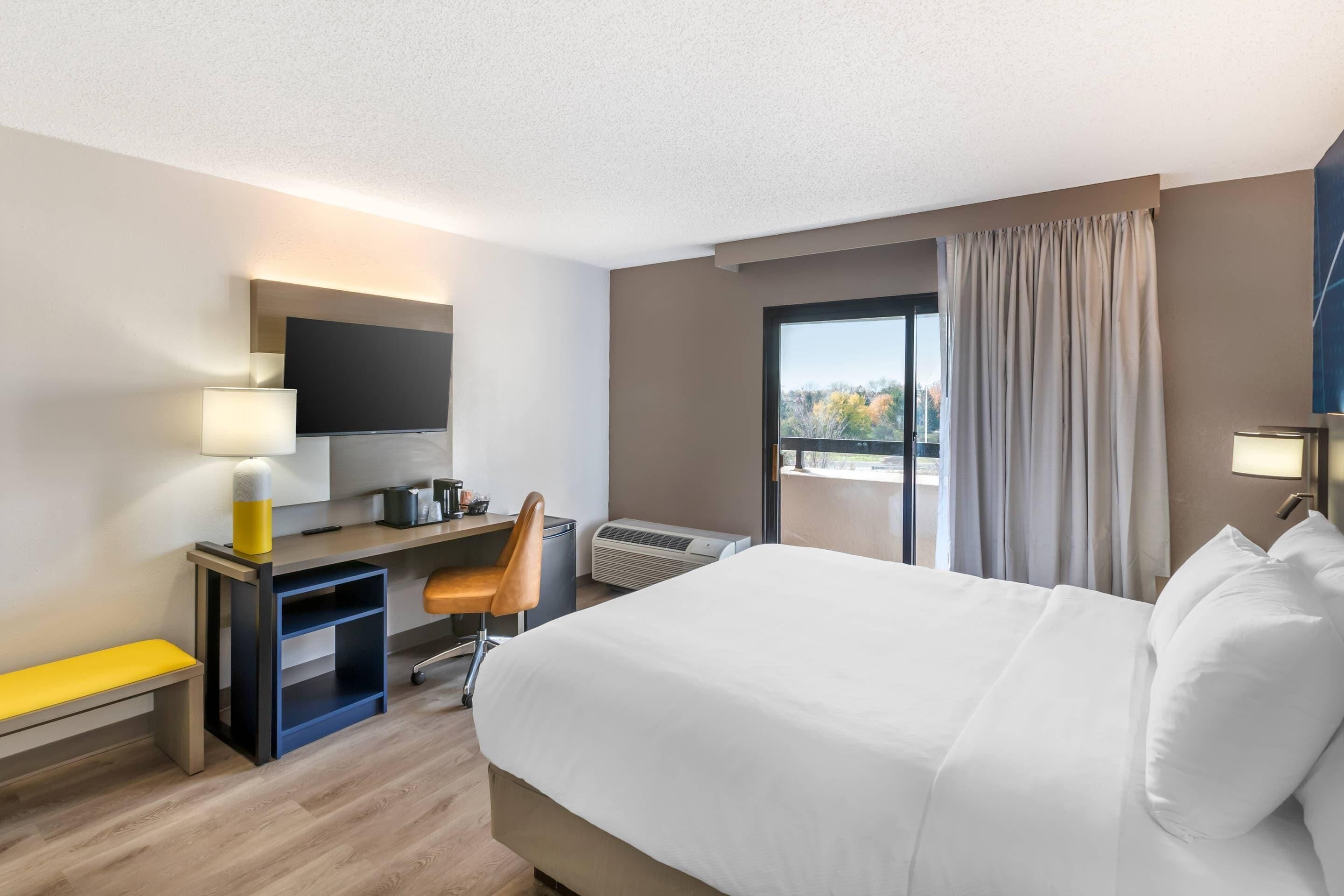 A hotel room with a bed, desk, chair, and a large window with curtains.