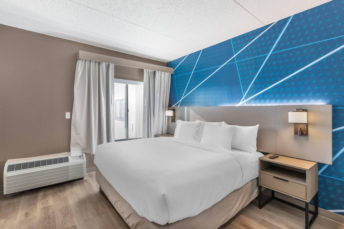 Una habitación de hotel moderna con una cama grande, una mesita de noche, una lámpara de pared y un ventanal con cortinas.