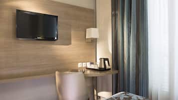 A modern hotel room with a flat-screen TV mounted on the wall, a small desk with a lamp, a chair, and a bed with a floral-patterned bedspread.