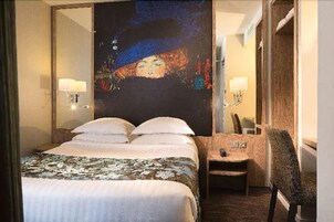 A hotel room with a large bed, a chair, a small table, and a painting on the wall.
