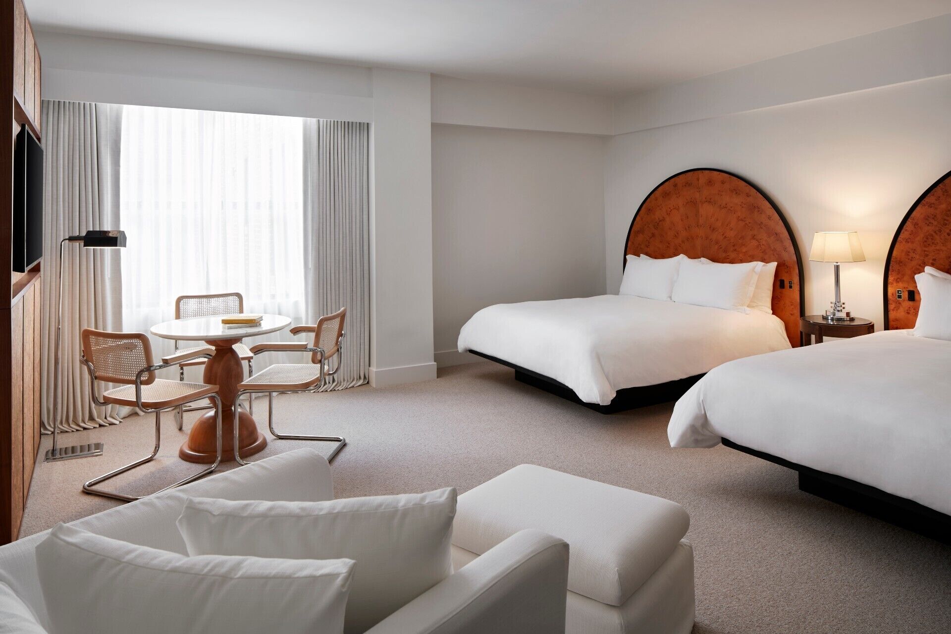 A hotel room with two beds, a desk with chairs, a TV, and a lamp.