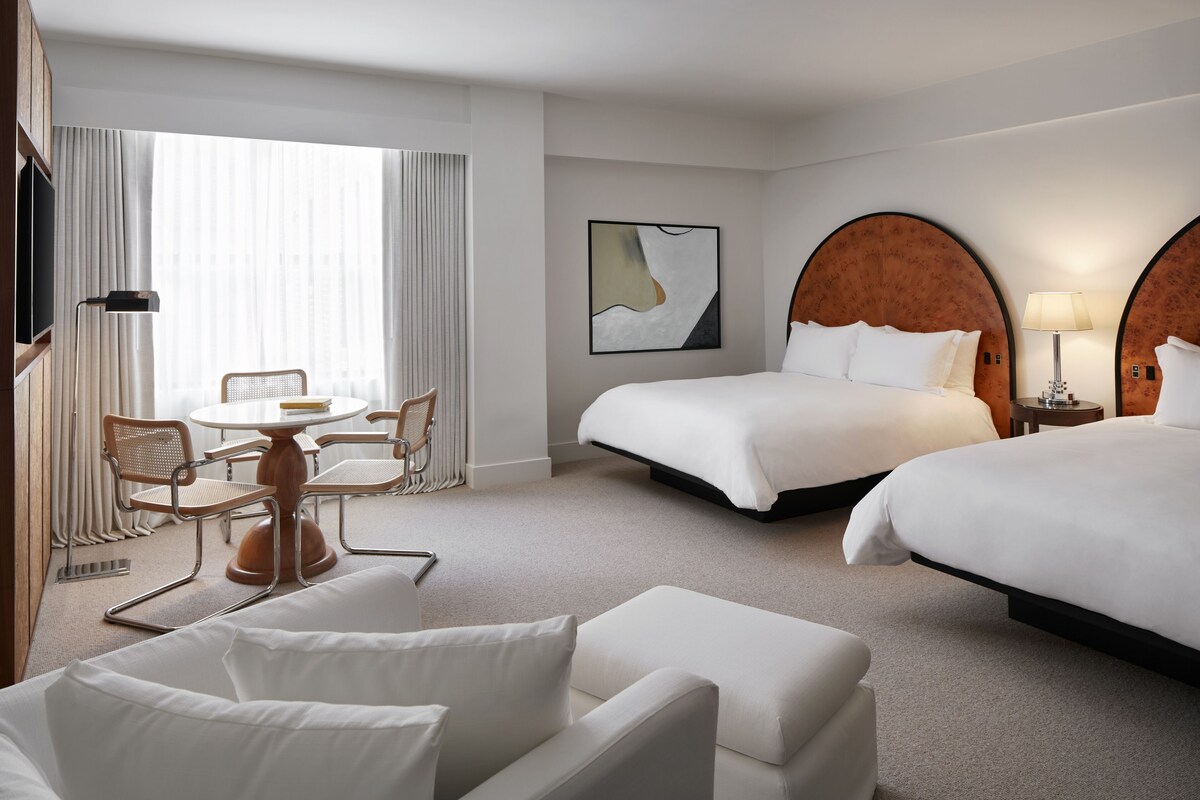 A hotel room with two beds, a dining area with a table and chairs, and a large window with curtains.