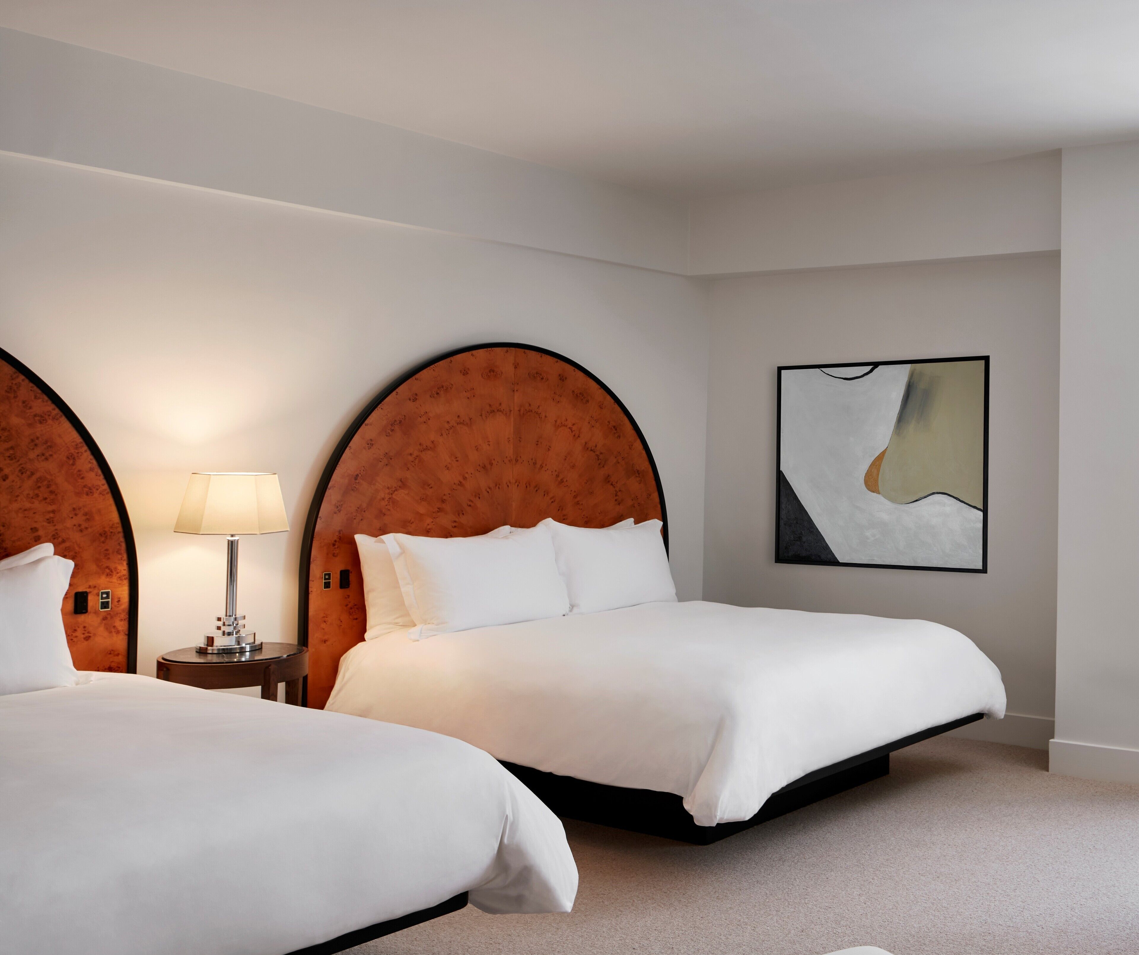A hotel room with two beds, a round wooden headboard, a bedside table with a lamp, and a modern abstract painting on the wall.