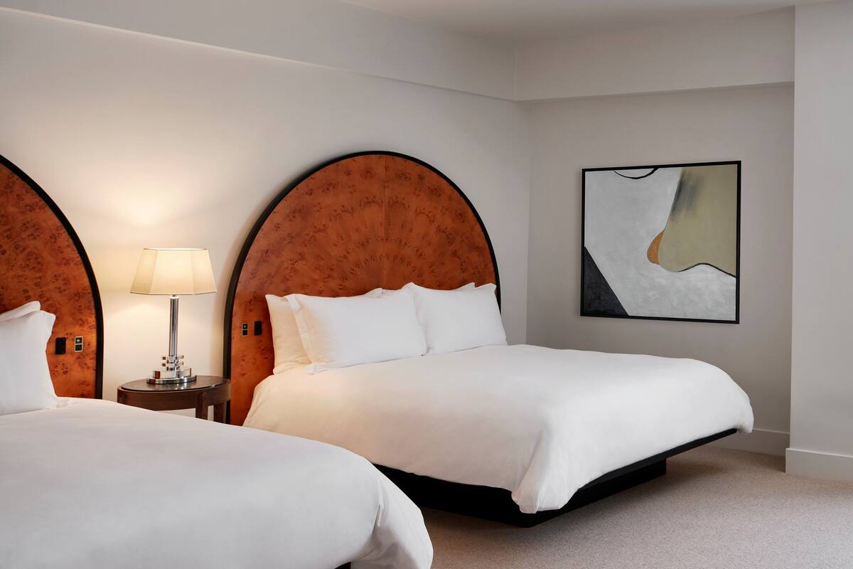 Habitación de hotel con dos camas, cabecero de madera redonda, mesita de noche con lámpara y un cuadro abstracto moderno en la pared.