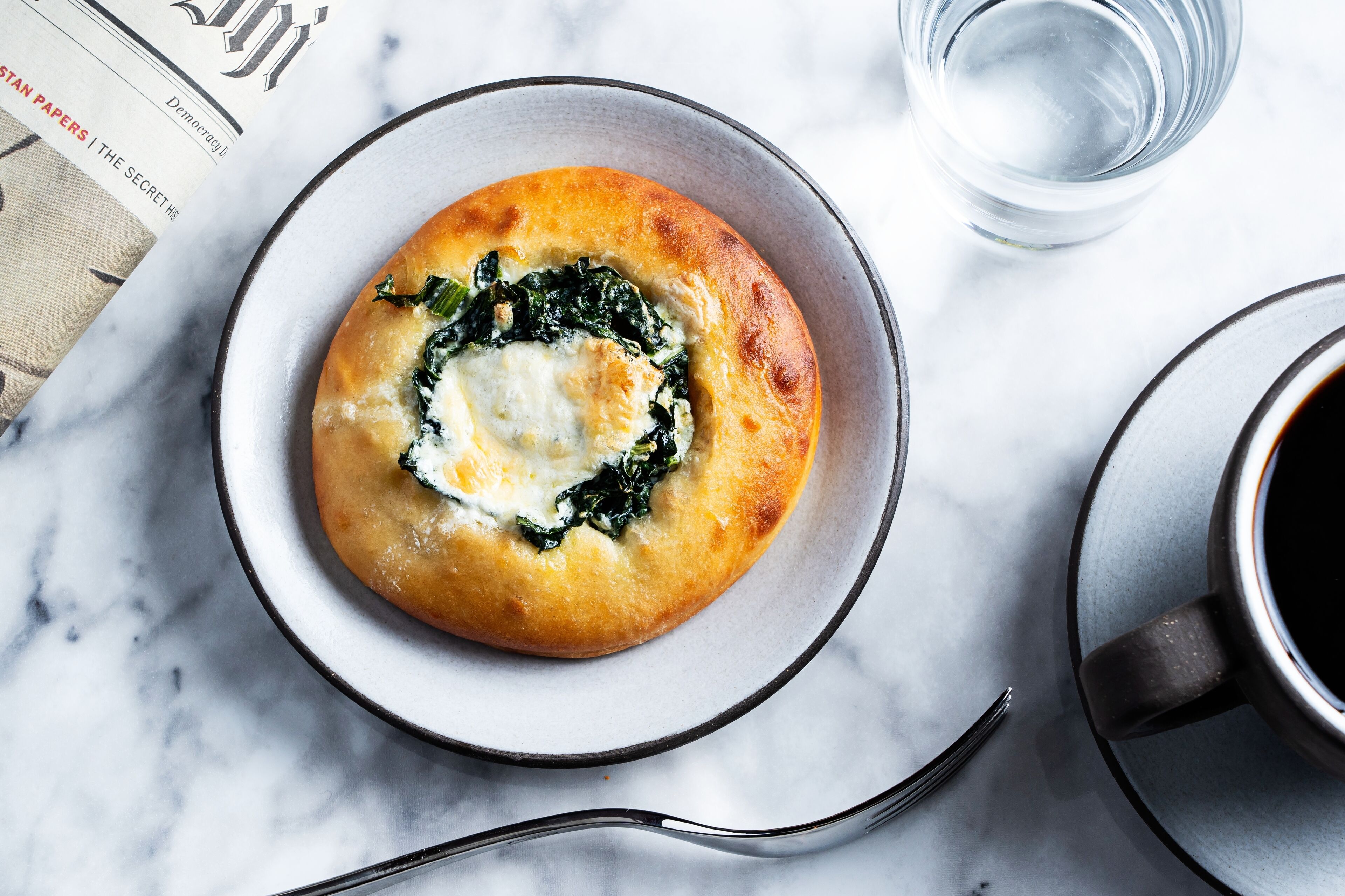 A bowl of spinach and cheese on a plate, a glass of water, and a cup of coffee on a marble surface.