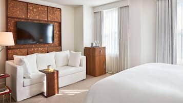 Ein Hotelzimmer mit einem großen Bett, einem weißen Sofa, einem Holzschrank, einem flachbildfernseher, der an einer strukturierten Wand angebracht ist, und einem großen Fenster mit Vorhängen.
