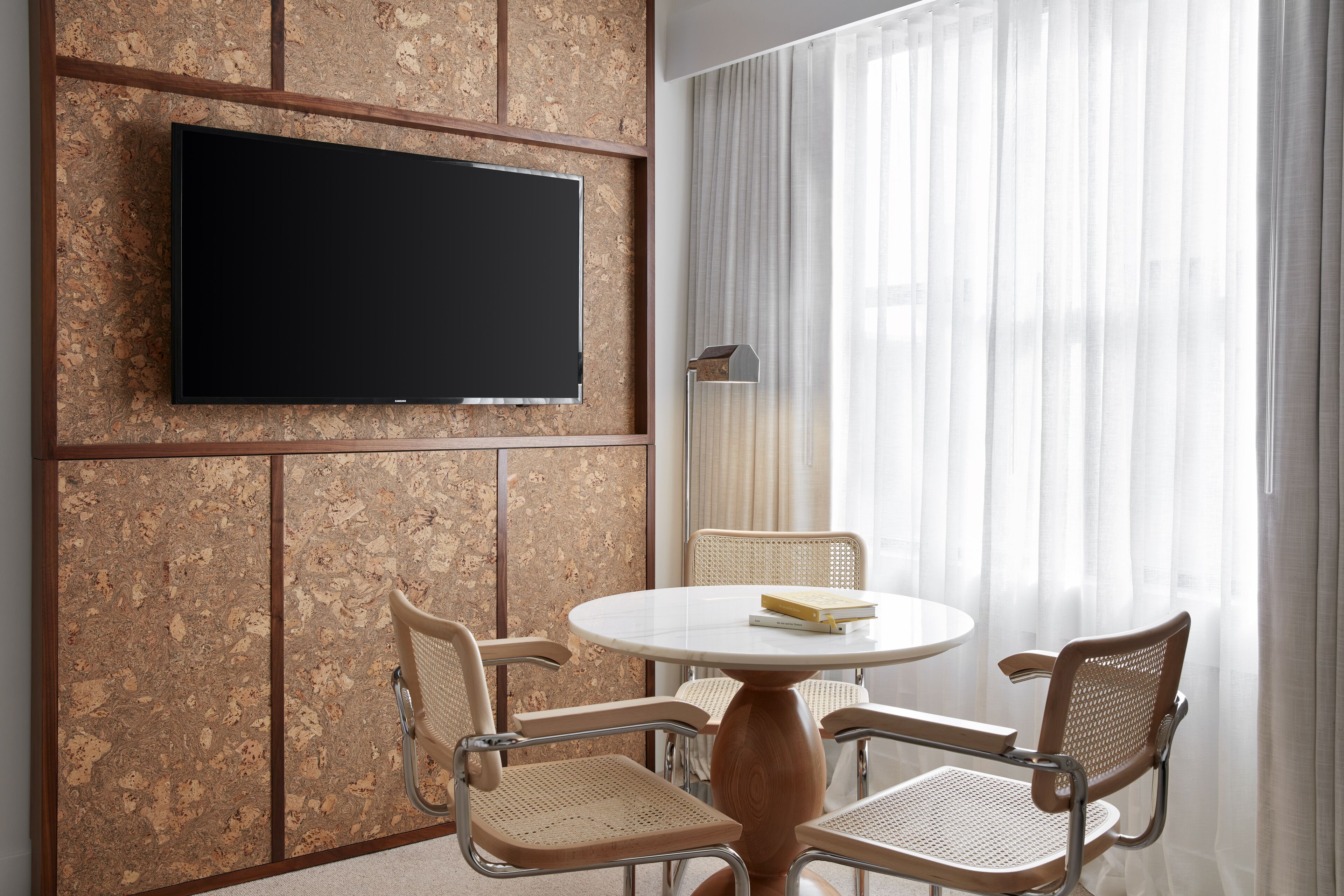 A modern room with a flat-screen TV mounted on a corkboard wall, a round table with two wicker chairs, and a large window with sheer curtains.