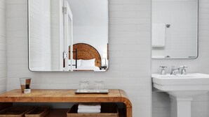 A modern bathroom with a wooden vanity, a white stool, a large mirror, and a white pedestal sink.