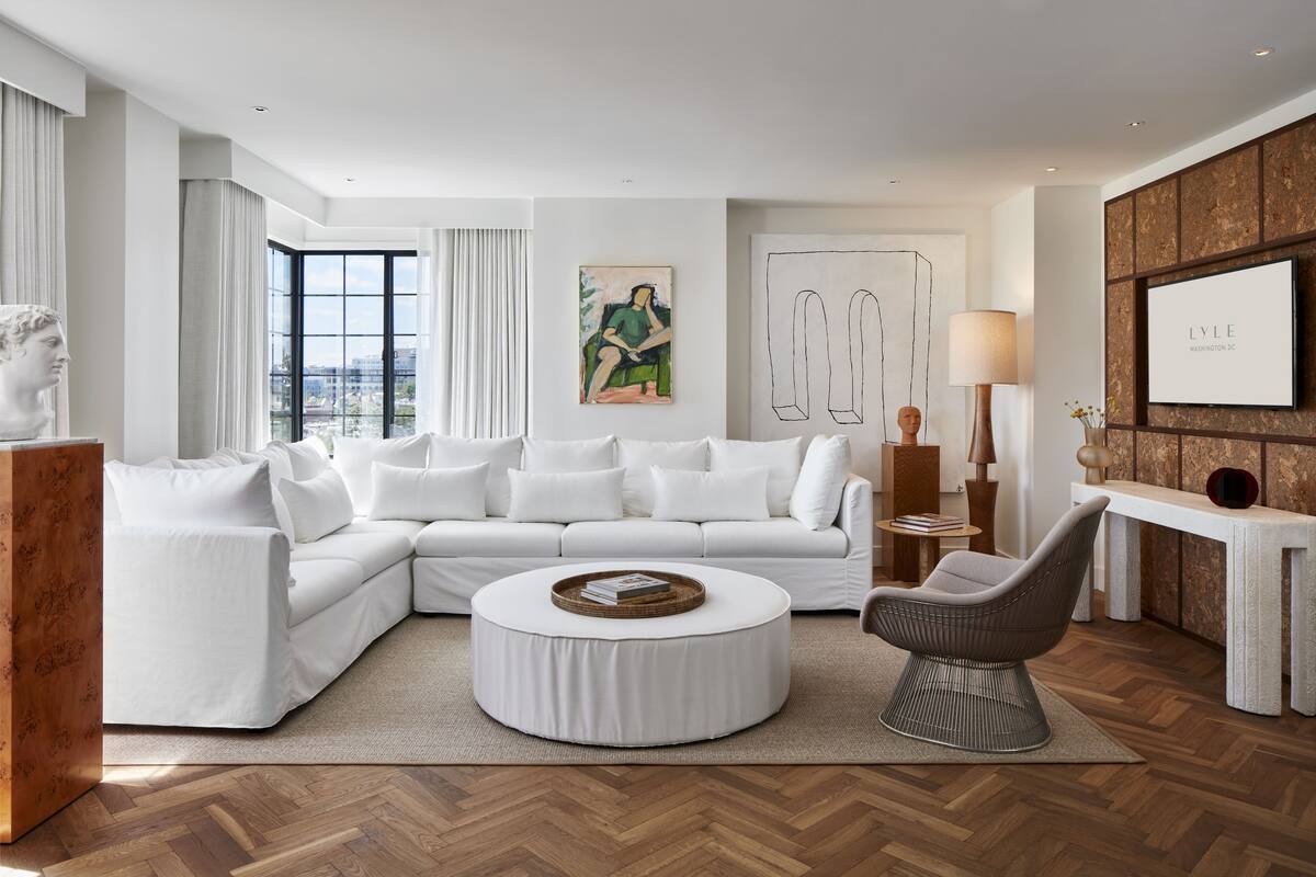 Una sala de estar moderna con un sofá seccional blanco, una mesa de centro redonda y un sillón gris. Se aprecia una ventana grande con vistas a la ciudad, un cuadro en la pared y una fotografía enmarcada sobre el sofá.