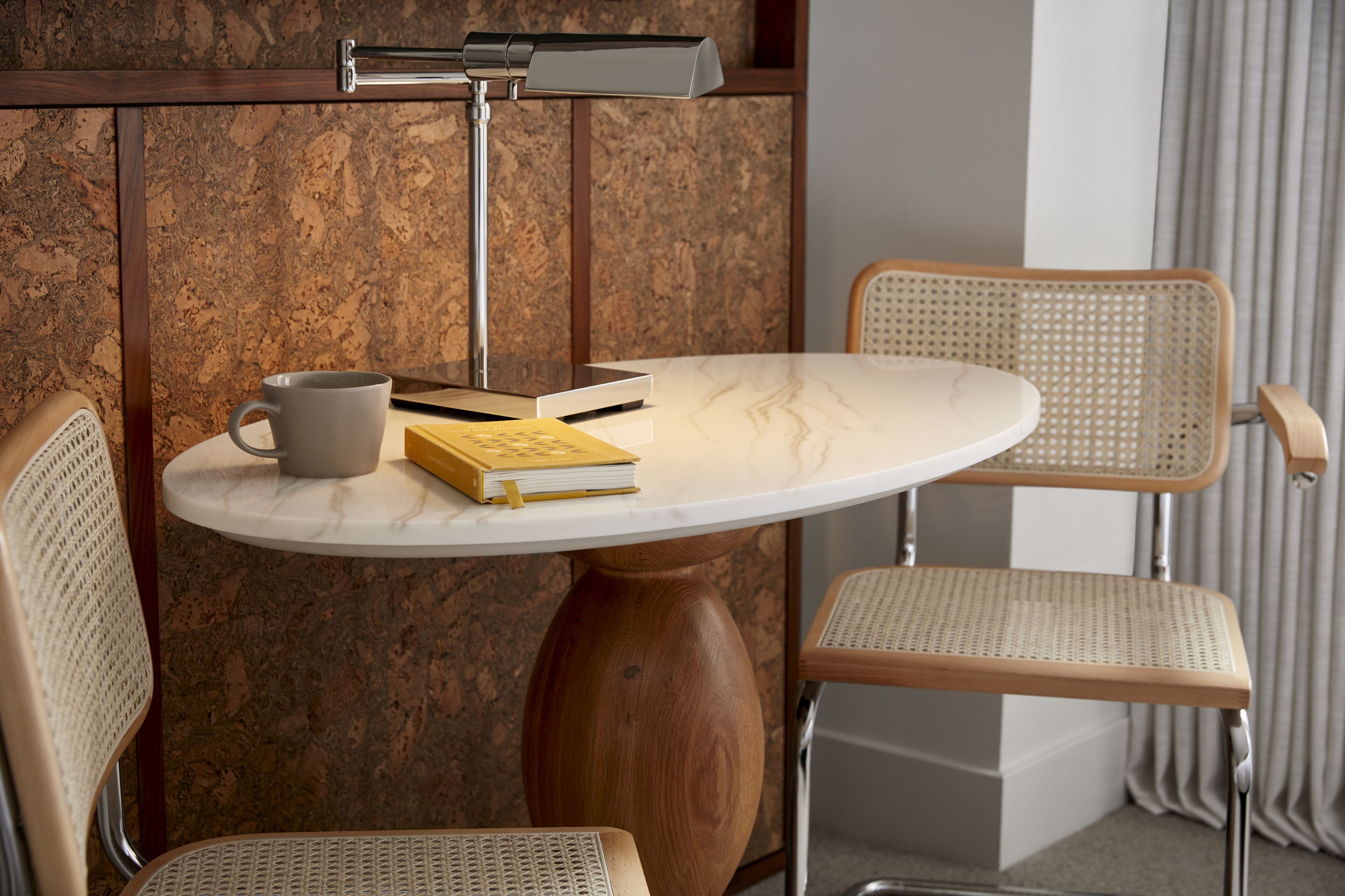 A round table with a white marble top, two wicker chairs, a grey mug, and a book on the table.