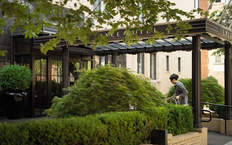 A hotel entrance with a covered walkway, greenery, and a person walking.