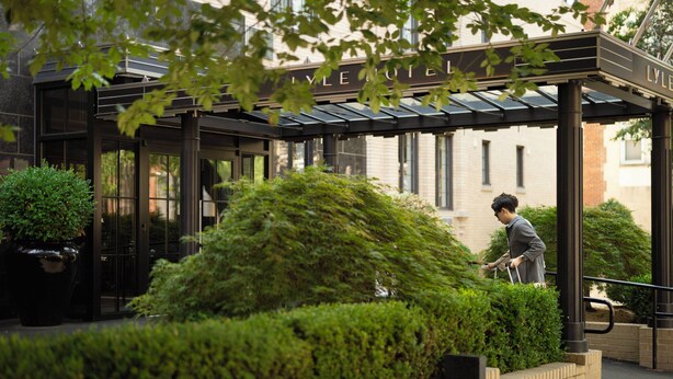 L’entrée d’un hôtel, avec une allée couverte, de la verdure et une personne qui marche.