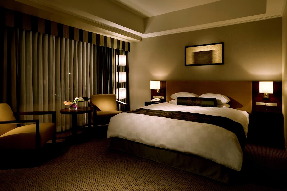 Habitación de hotel con una cama grande, dos mesitas de noche con lámparas, una silla, una mesita pequeña con un florero y una ventana con cortinas.
