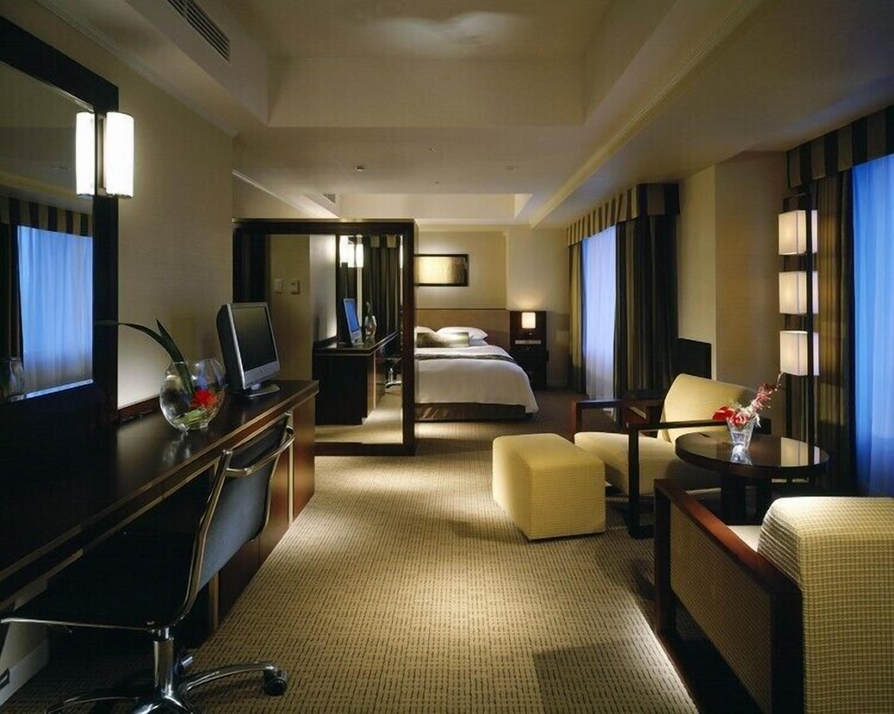 A hotel room with a bed, a desk with a computer, a chair, a small table with a vase, and a mirror.