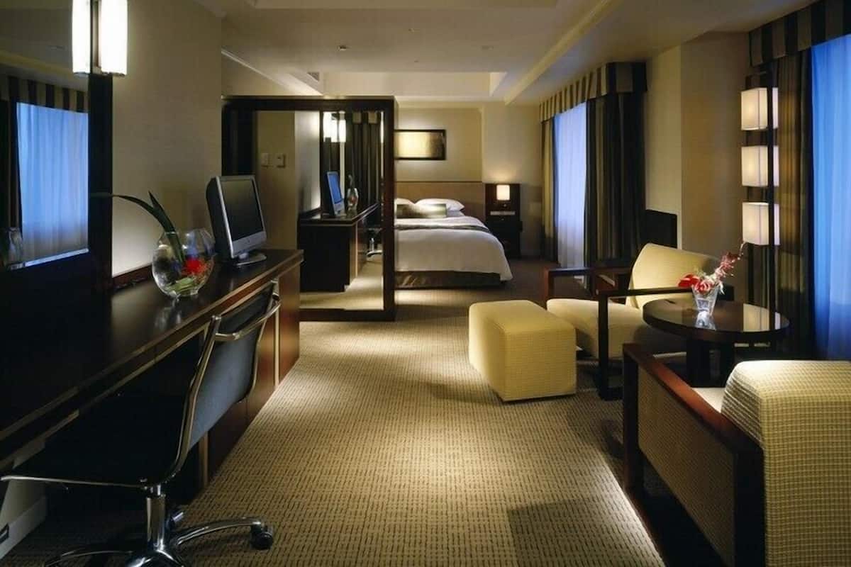 Habitación de hotel con cama, escritorio con computadora, silla, una mesita con un florero y un espejo.