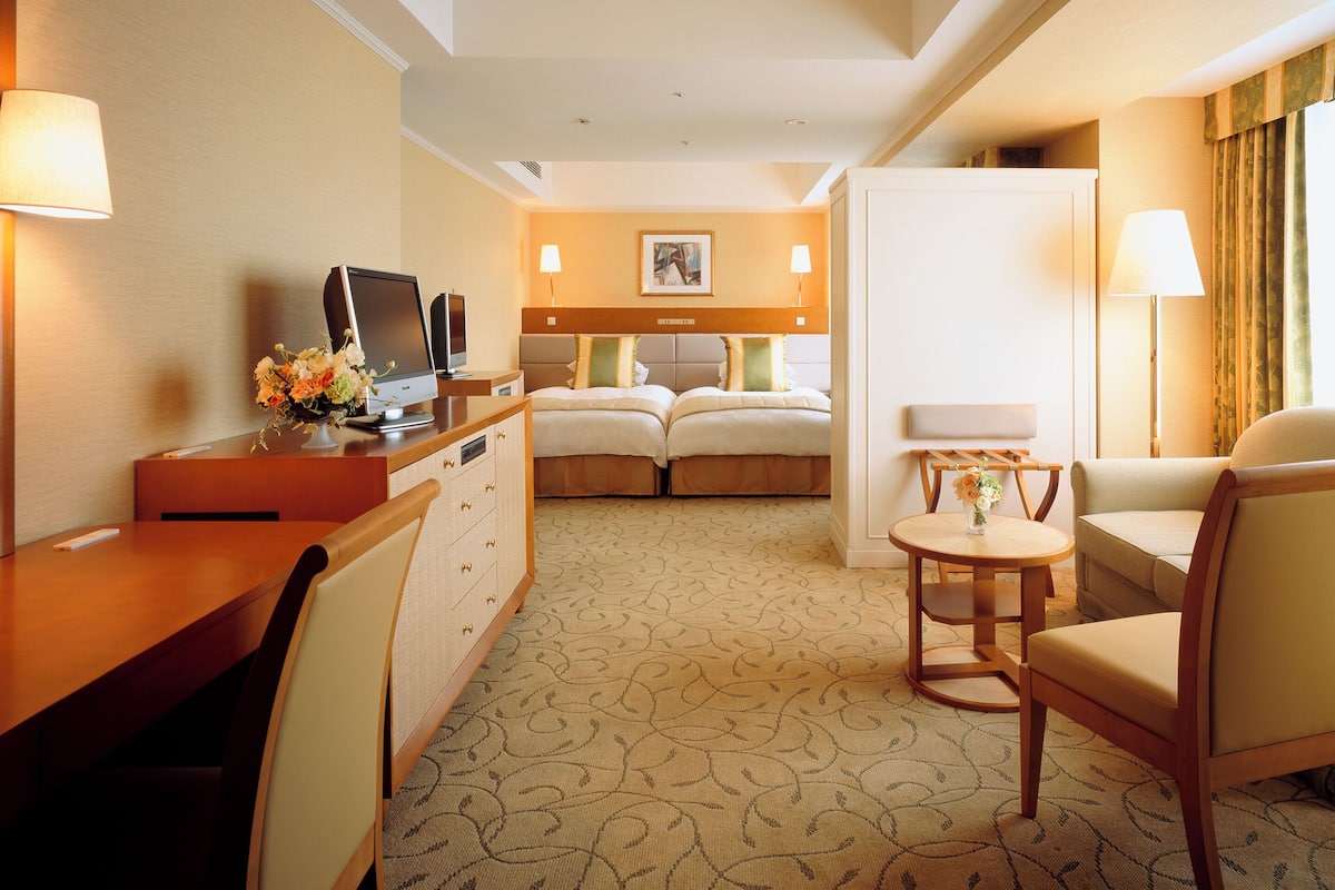Una habitación de hotel con escritorio, dos camas, zona de estar y una mesita con lámpara.