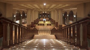 Una imponente escalera conduce a un vestíbulo con un arreglo floral central, flanqueado por columnas y barandillas de madera.