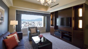 Habitación de hotel con un televisor grande, un sofá, una mesa de centro con flores y vistas a la ciudad.