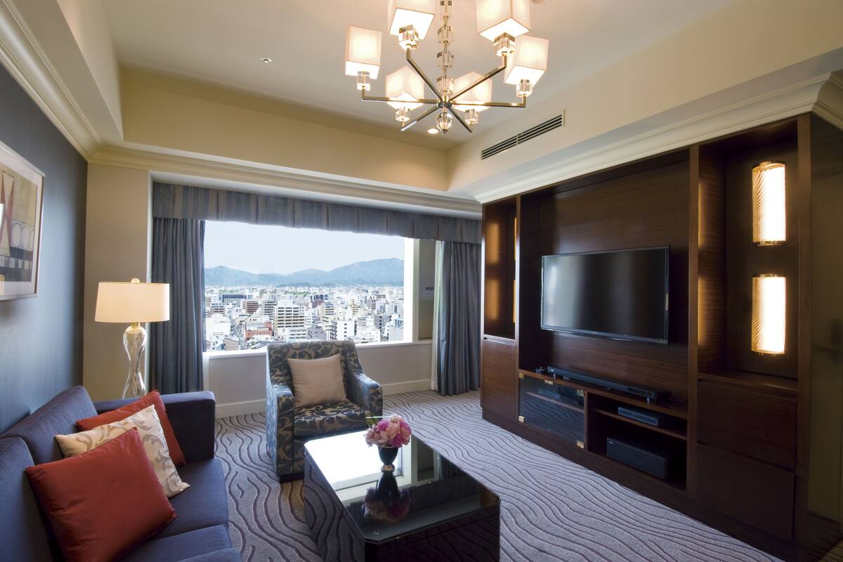 Habitación de hotel con un televisor grande, un sofá, una mesa de centro con flores y vistas a la ciudad.