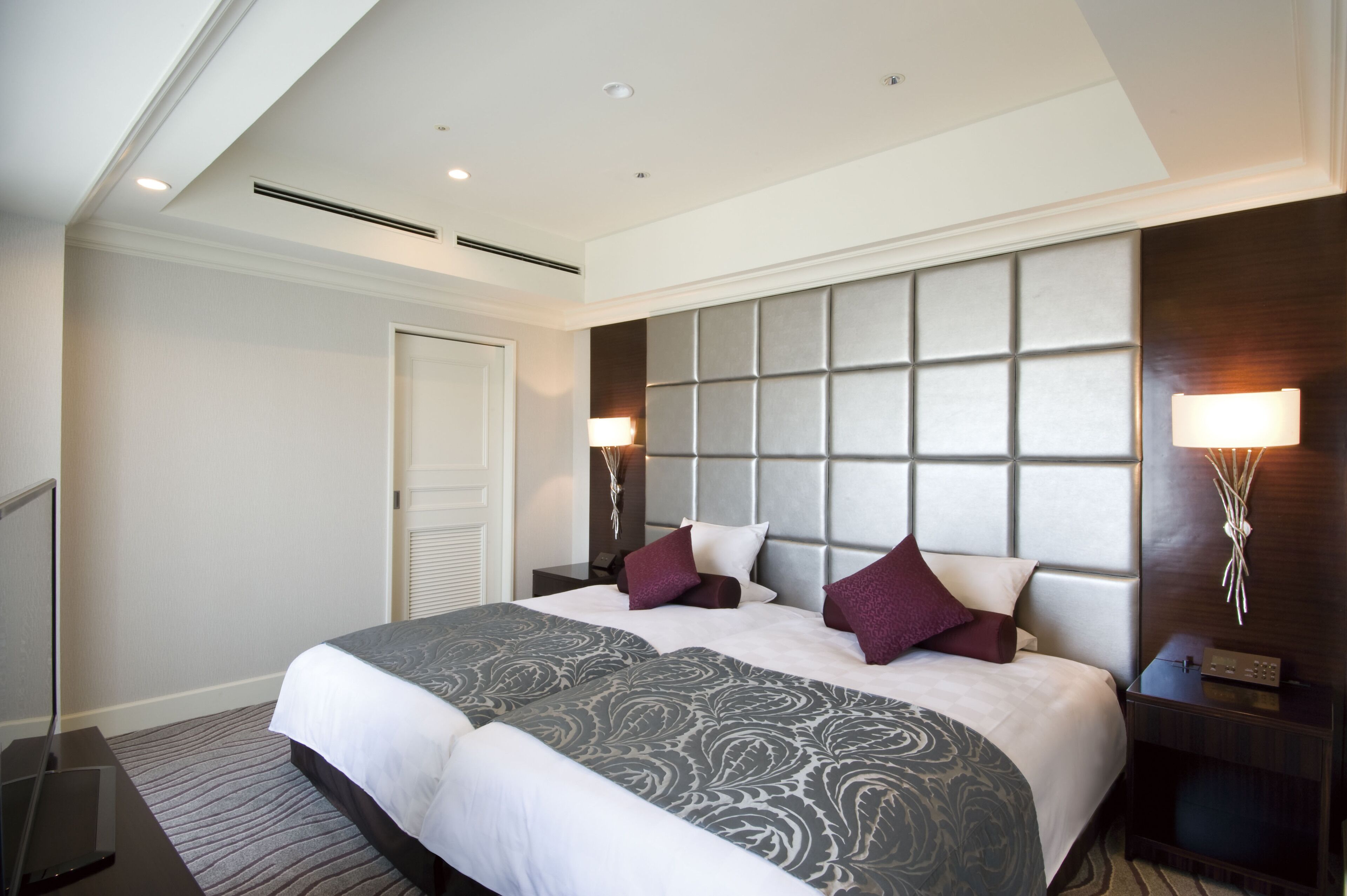 A modern hotel room with a large bed, a headboard with a padded panel, two bedside lamps, and a flat-screen TV on a stand.