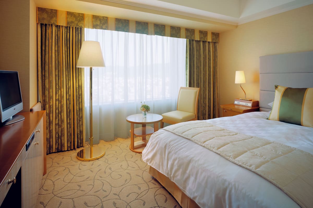 Una habitación de hotel con una cama, un escritorio con computadora, una silla, una mesita con lámpara y una mesita de noche con lámpara.