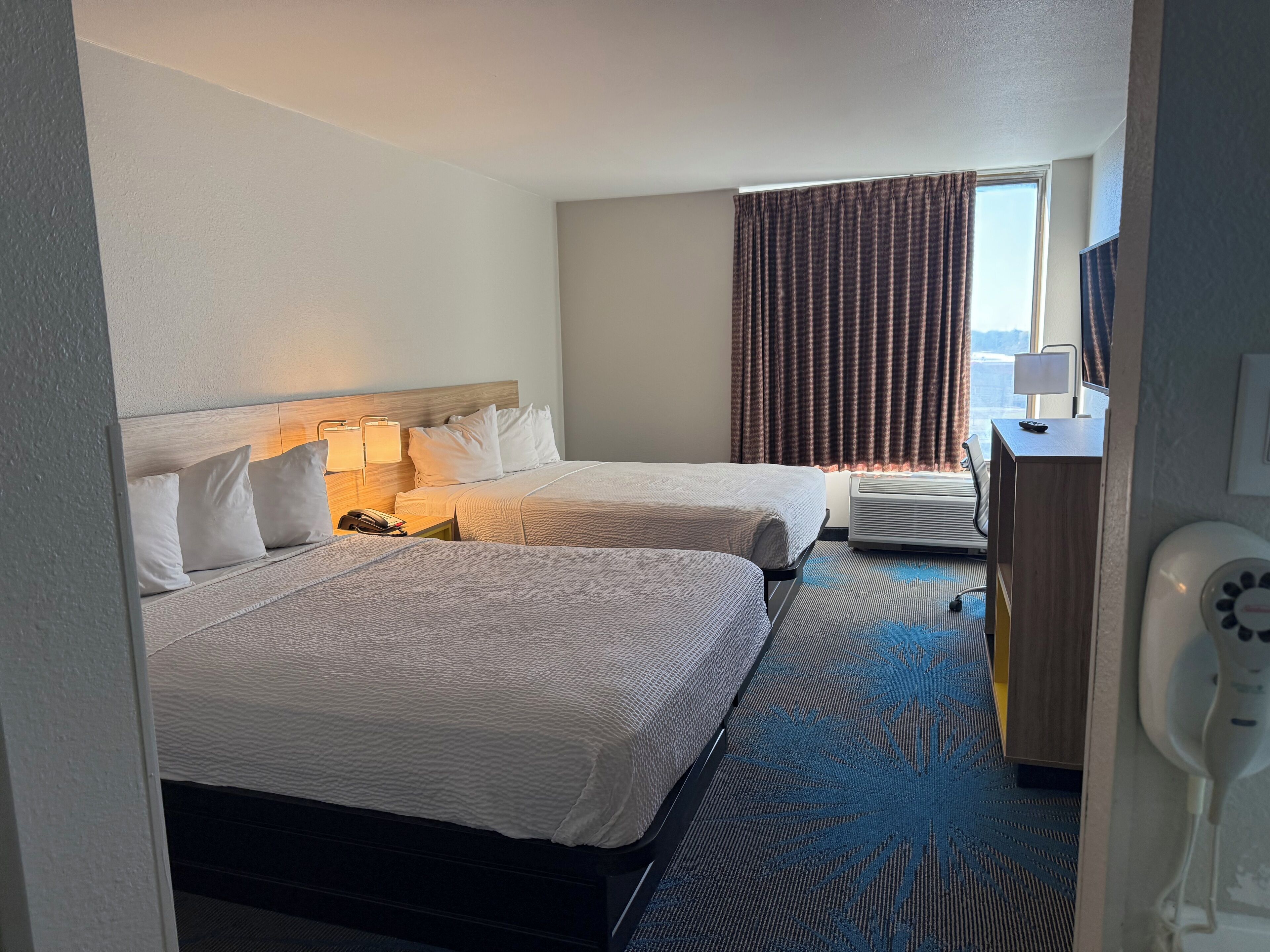 Une chambre d’hôtel avec deux lits, un bureau, une chaise, une lampe et une fenêtre avec des rideaux.