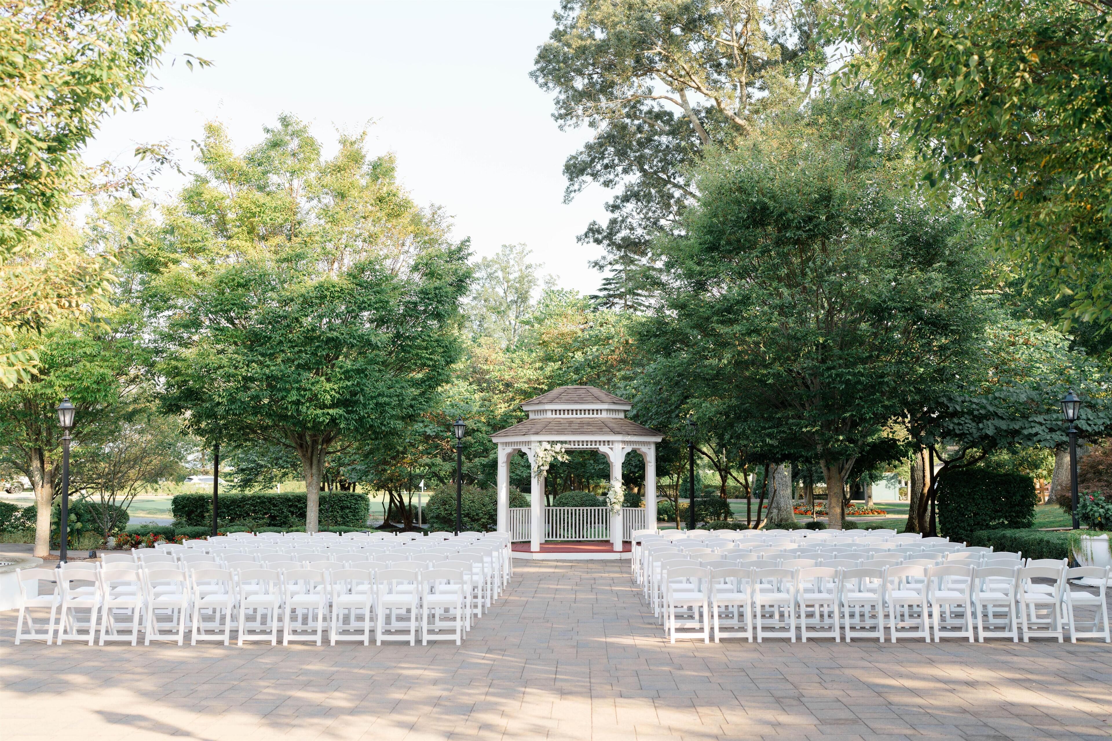 A gazebo with white chairs arranged in rows, surrounded by trees and a brick pathway.
