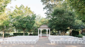 A gazebo with white chairs arranged in rows, surrounded by trees and a brick pathway.
