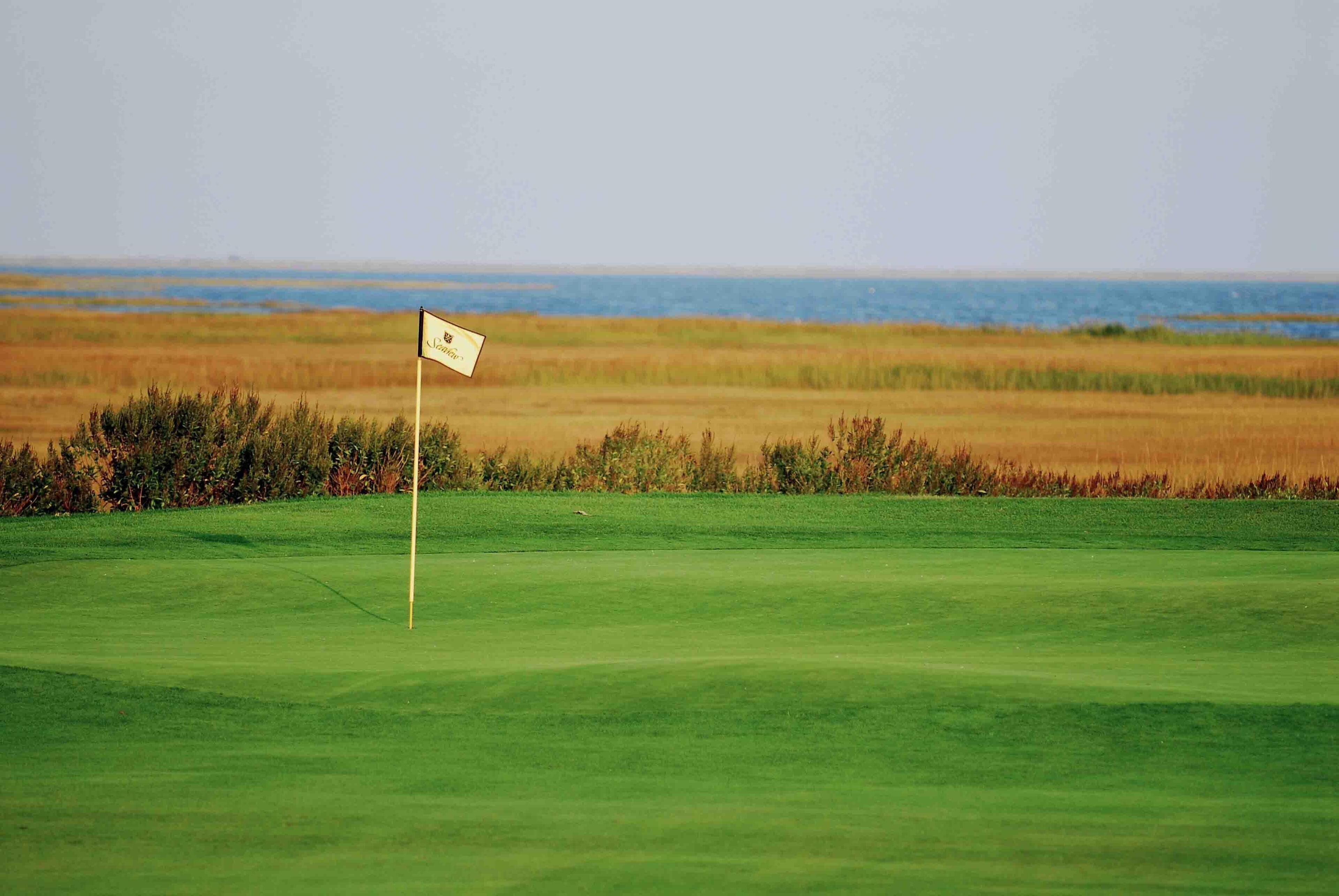 A golf course with a flag on the green, a clear sky, and a distant view of the ocean.