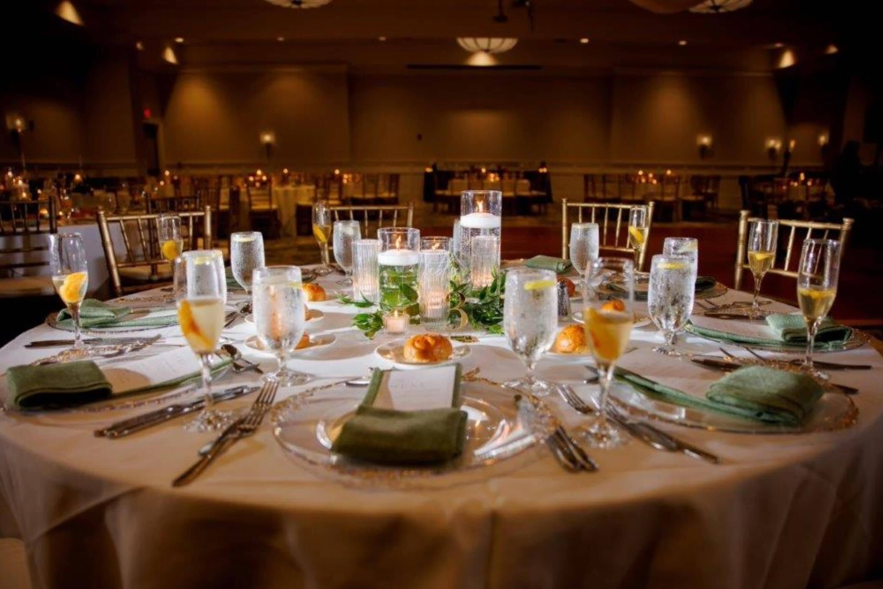 Una mesa de banquete dispuesta con varios vasos, platos y cubiertos, rodeada de sillas.