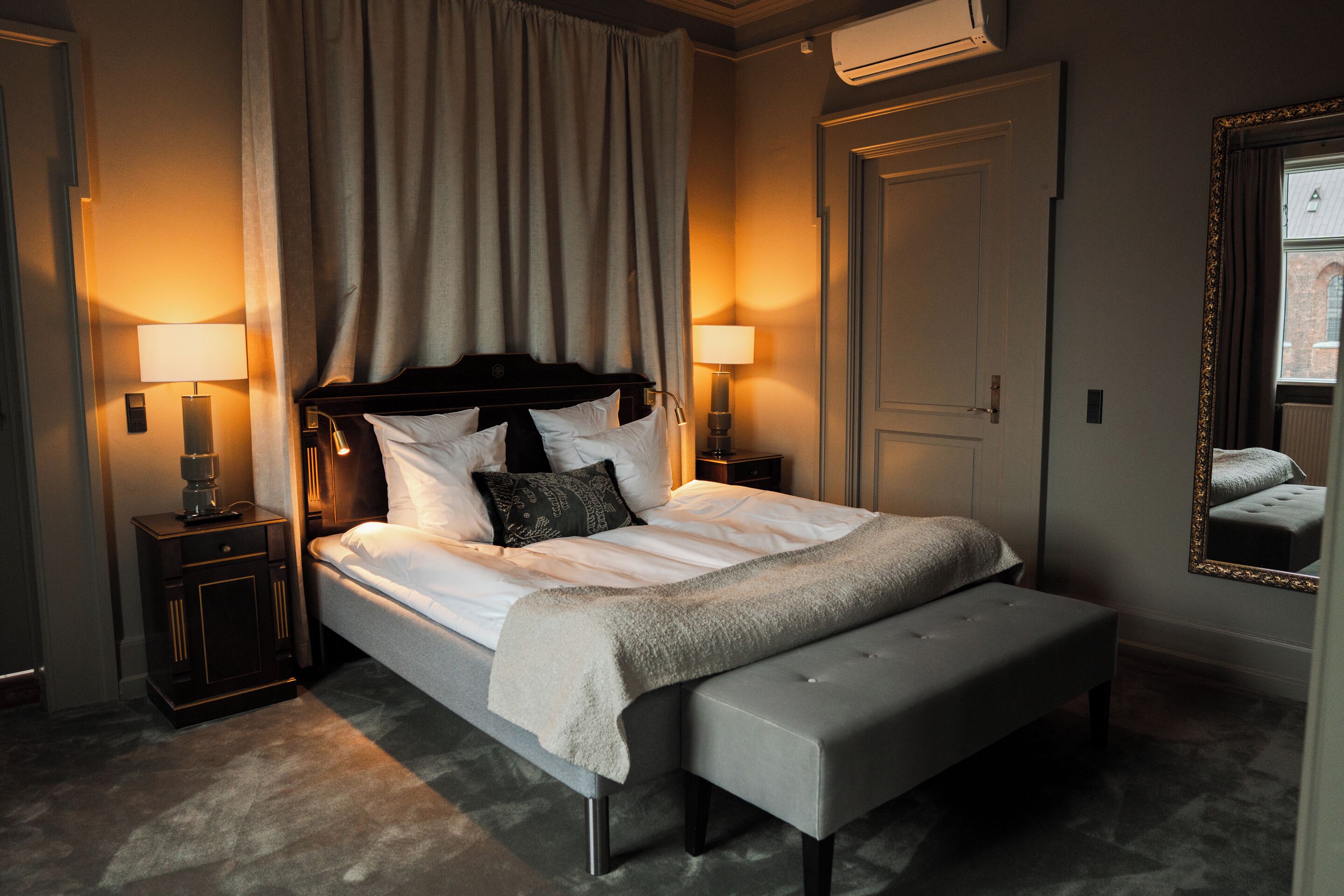A hotel room with a large bed, two bedside tables with lamps, a bench, a mirror, and an air conditioning unit.