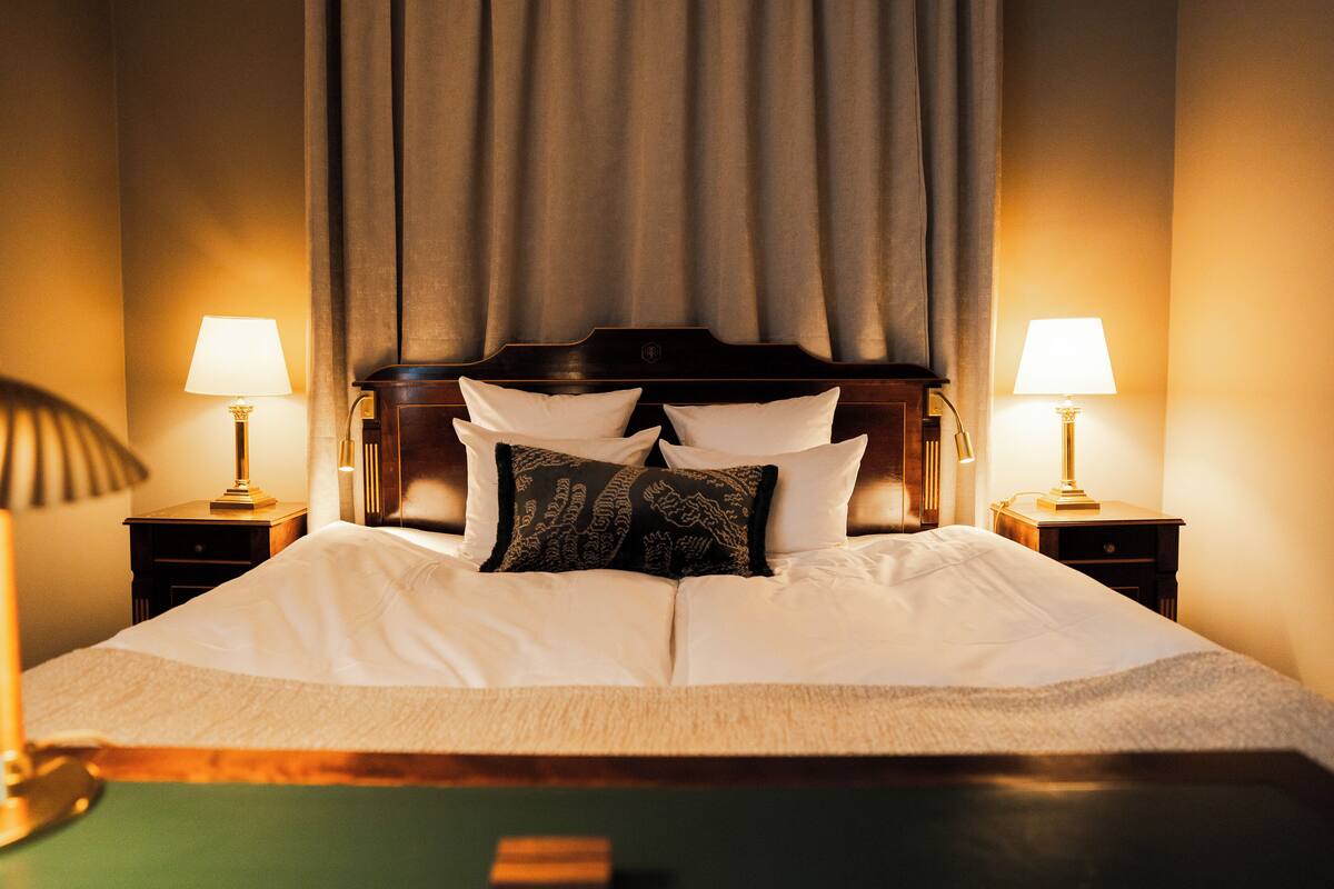 A neatly made bed with white linens and decorative pillows, flanked by two bedside tables with lamps, in a room with a large window and a curtain.