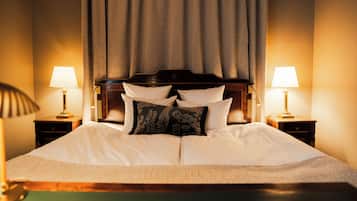 Ein ordentlich bezogenes Bett mit weißen Leinen und dekorativen Kissen, an beiden Seiten zwei Nachttische mit Lampen, in einem Zimmer mit großem Fenster und Vorhang.