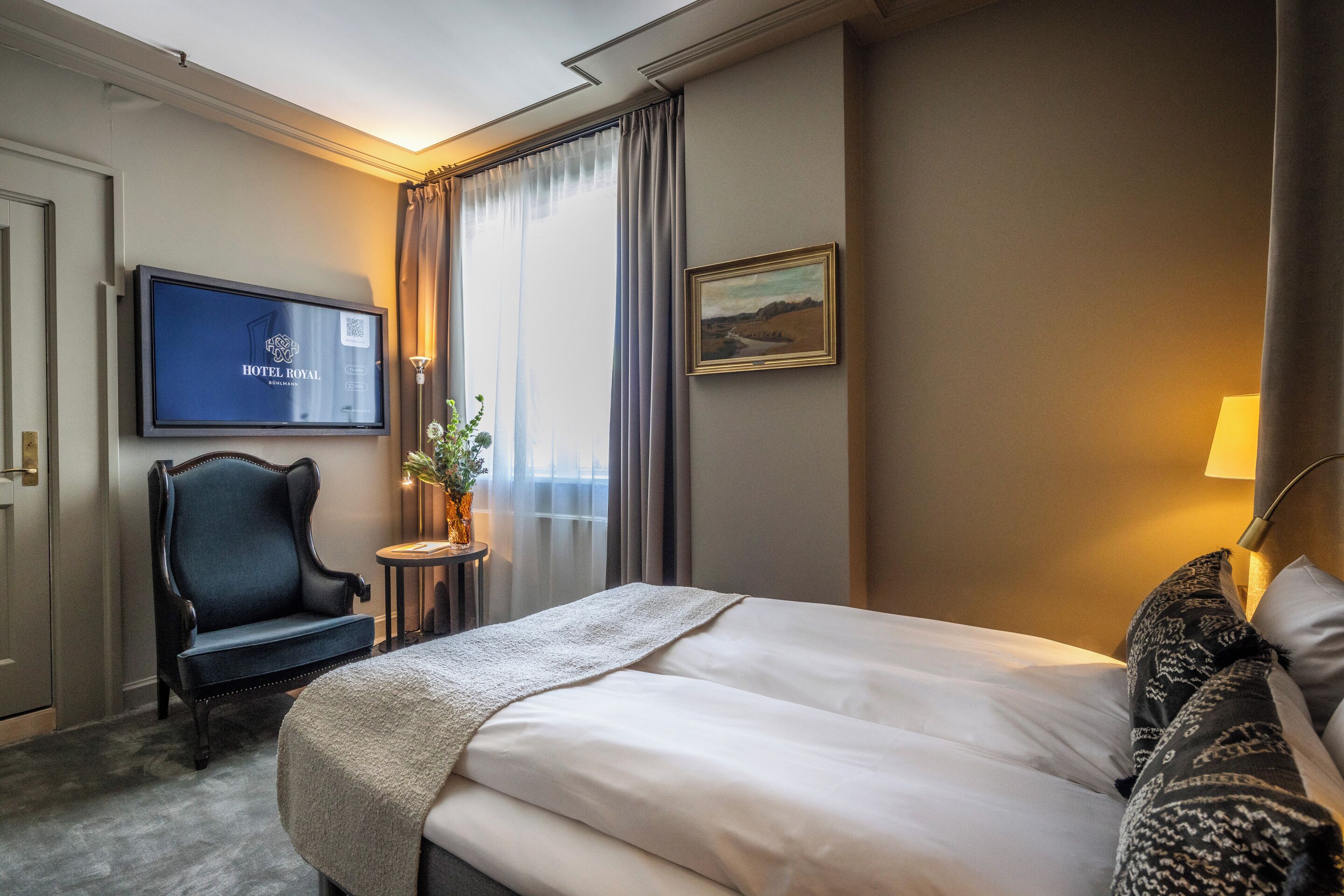 A hotel room with a bed, a TV mounted on the wall, a chair, a small table with a plant, and a painting on the wall.