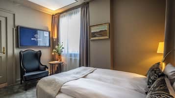 A hotel room with a bed, a TV mounted on the wall, a chair, a small table with a plant, and a painting on the wall.