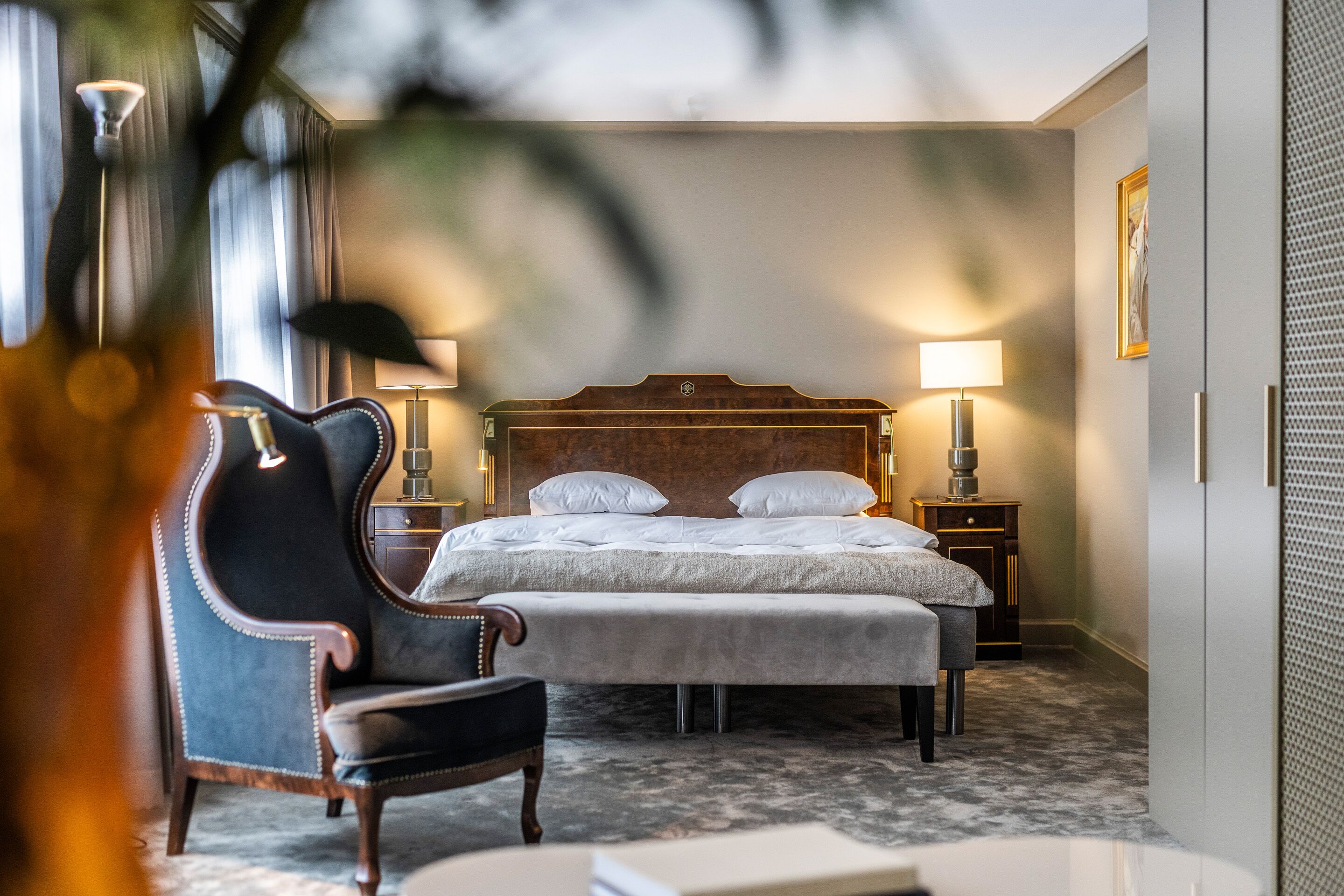 Quarto de hotel com uma cama grande, duas mesas de cabeceira com abajures, uma poltrona estofada escura e uma pequena mesa com luminária.